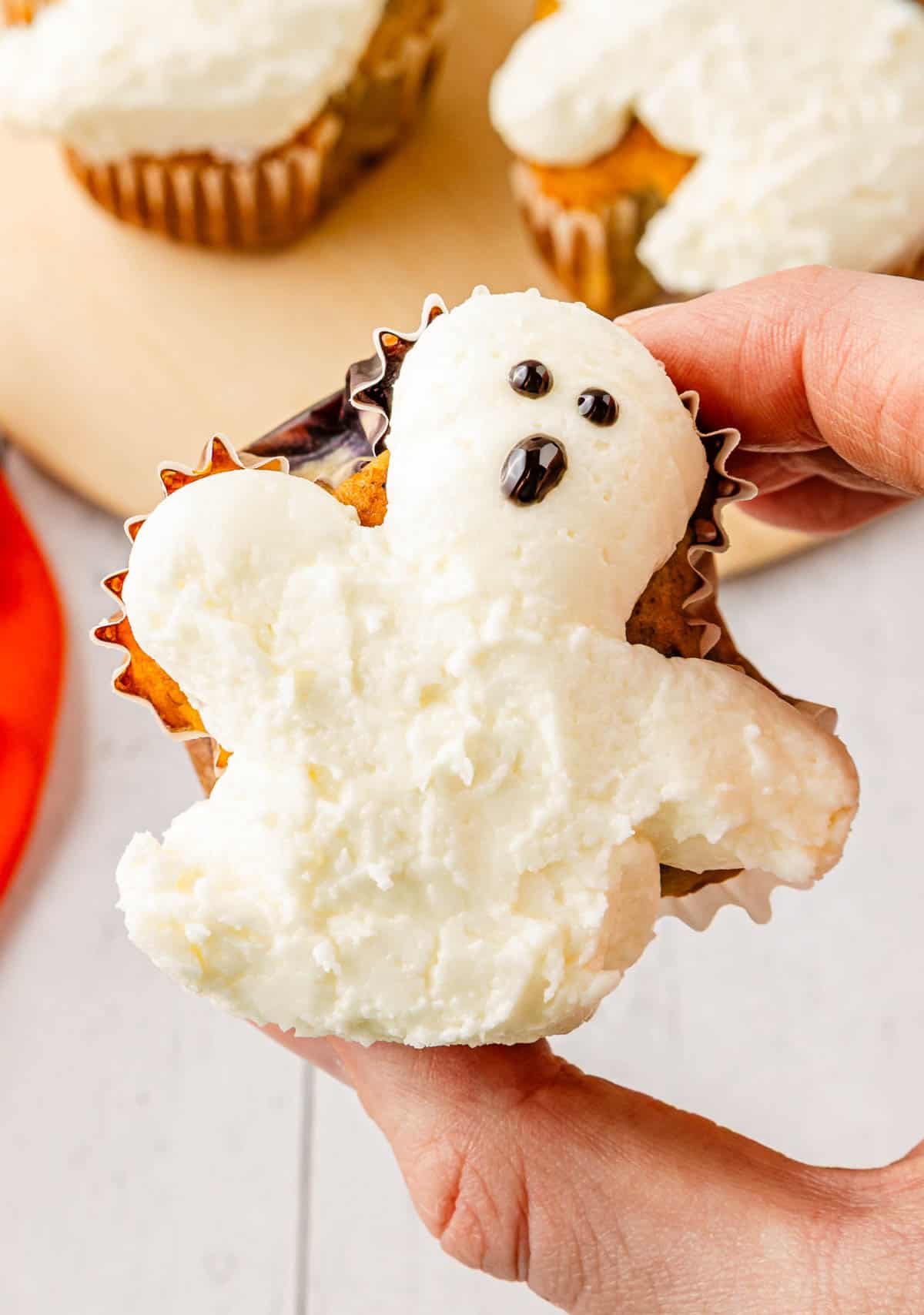 A hand holds a Ghost Muffin decorated with white cream cheese icing to look like a ghost, complete with chocolate eyes and mouth.