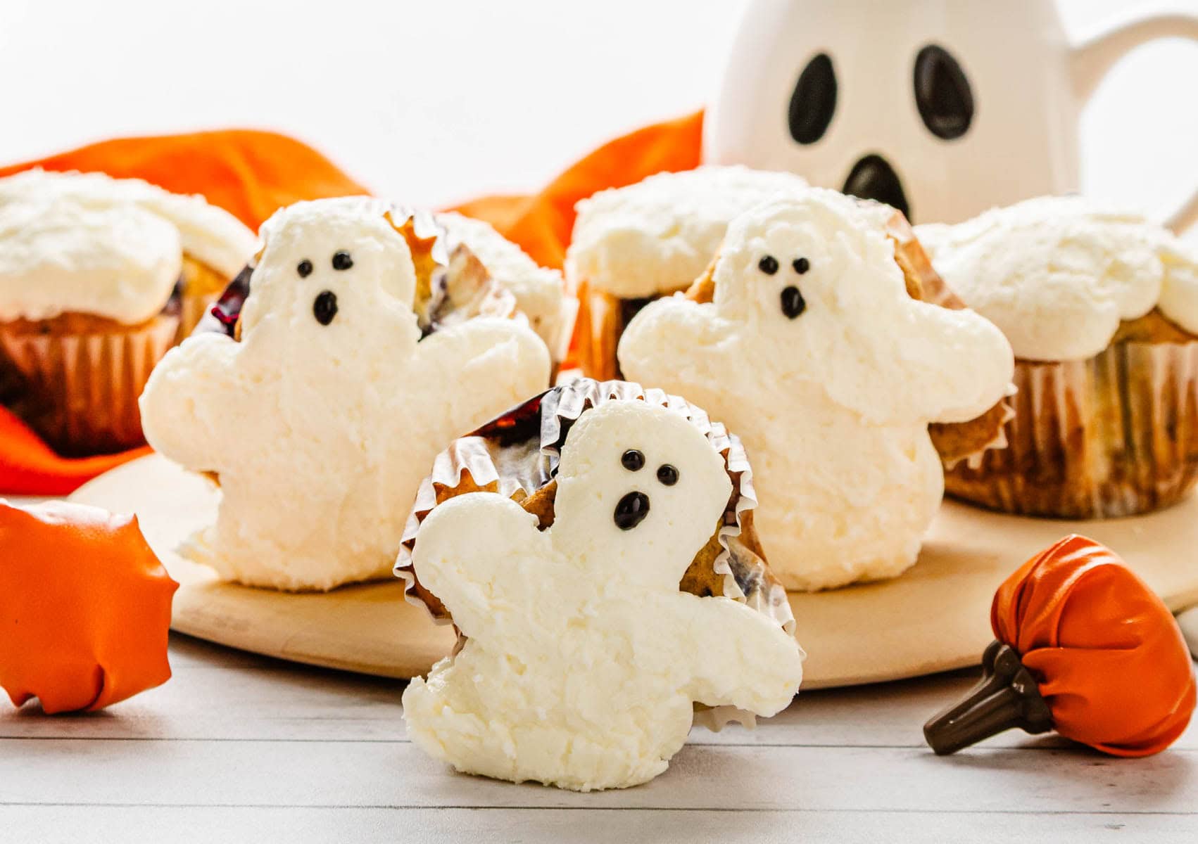 Cupcakes decorated as white ghosts with black eyes and mouths, surrounded by small pumpkin decorations. Try this BOO-berry Ghost Muffins recipe topped with spooky SCREAM Cheese Icing for a festive Halloween treat!.