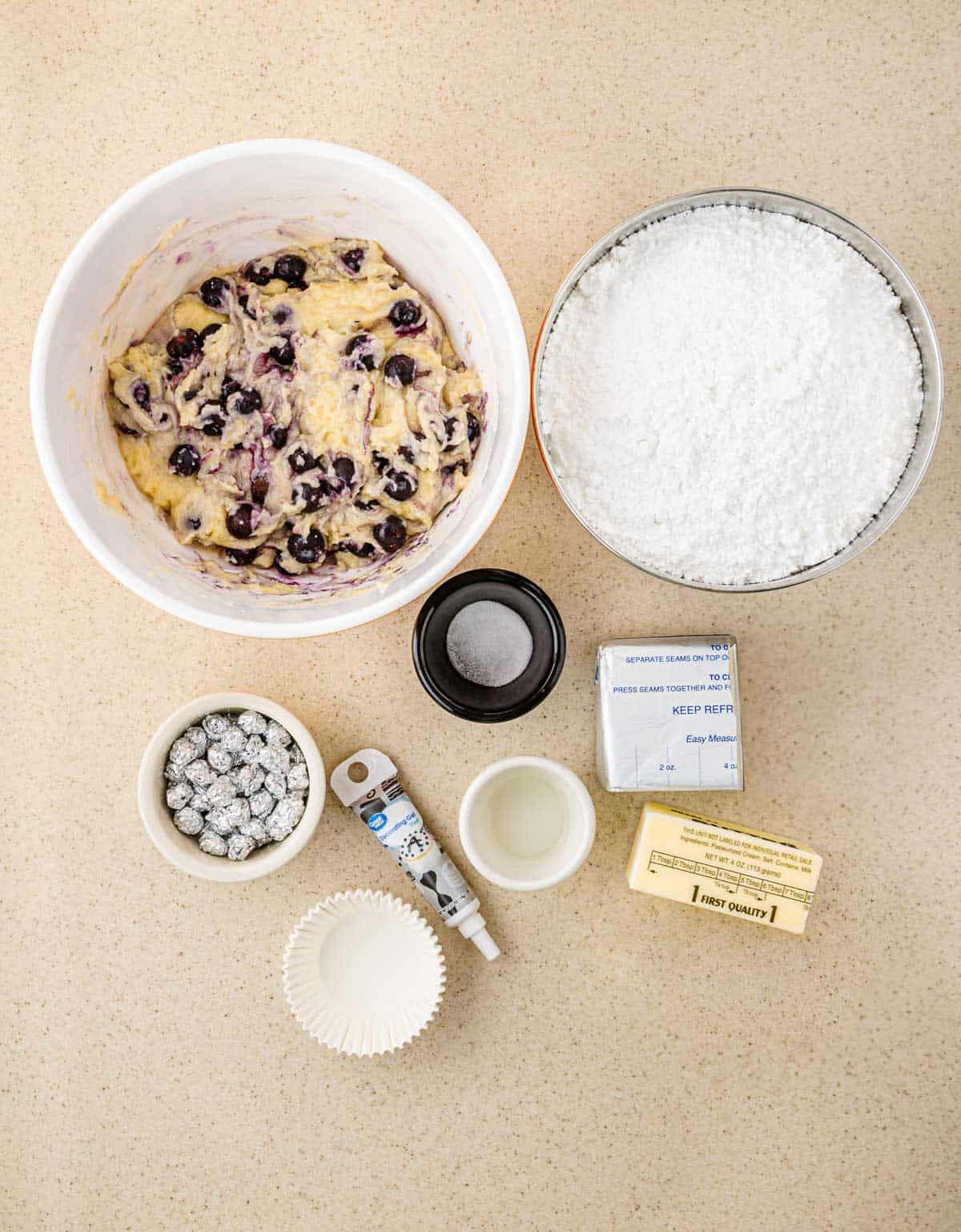 Bowls of BOO-berry batter and powdered sugar sit alongside butter, eggs, cupcake liners, and baking ingredients&mdash;everything you need to whip up ghost muffins topped with cream cheese icing.