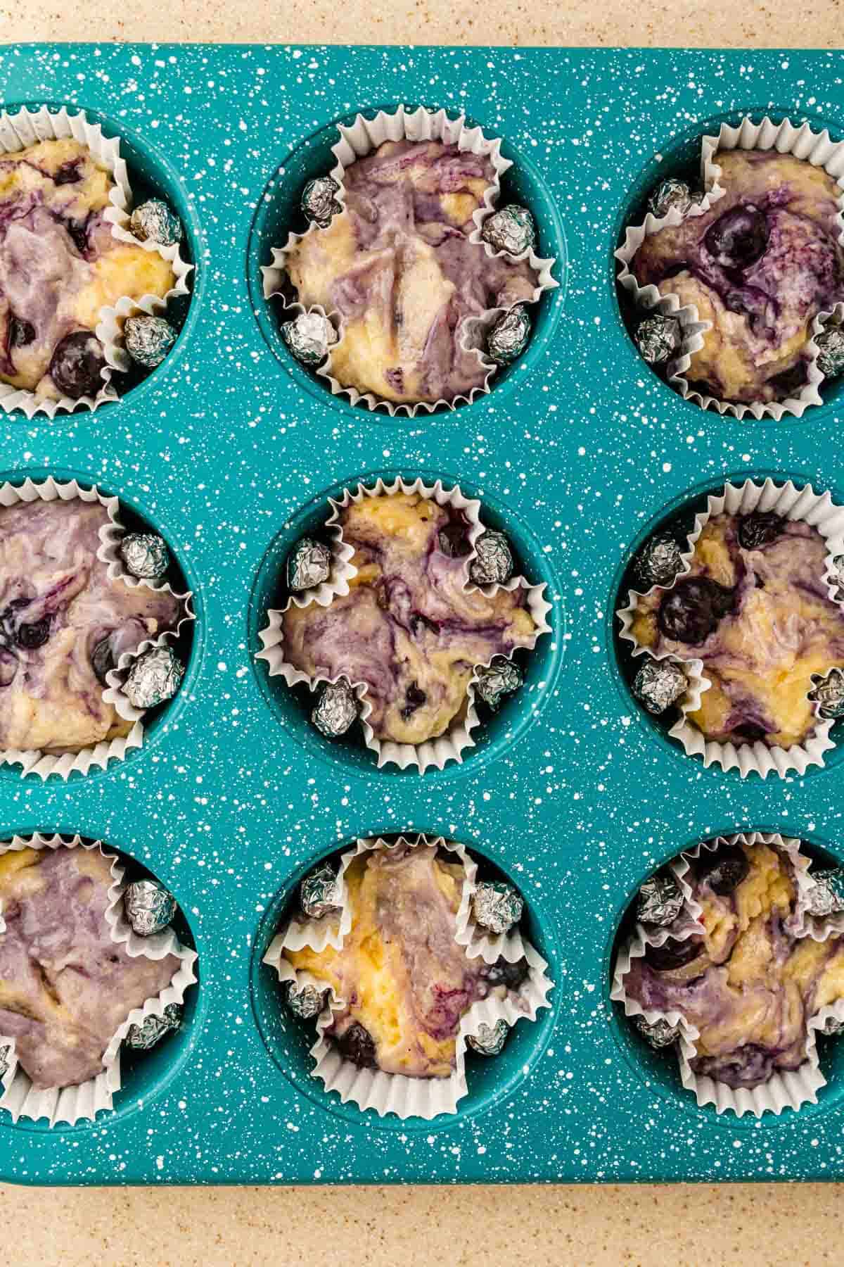 A teal muffin pan holds nine unbaked BOO-berry muffin batter portions in paper liners, ready to become spooky Ghost Muffins with Scream Cheese Icing.