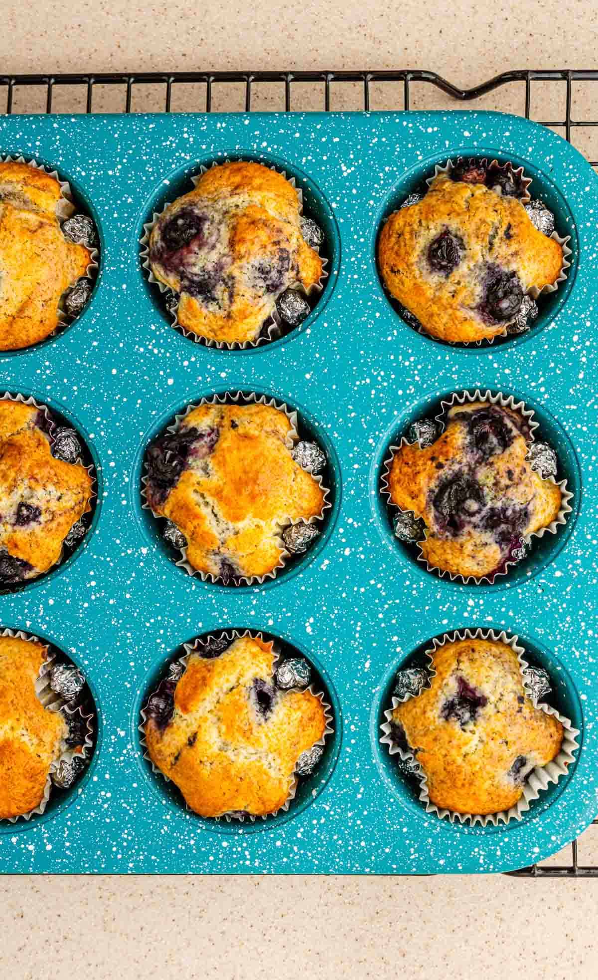 A teal muffin tin holds nine freshly baked BOO-berry Ghost Muffins on a cooling rack, perfect for a fun Halloween recipe.