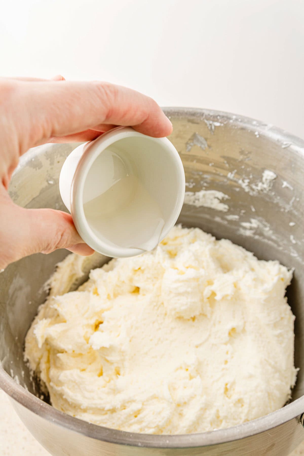 A hand pours liquid from a small cup into a mixing bowl filled with creamy BOO-berry frosting, perfect for topping Ghost Muffins or swirling into homemade Cream Cheese Icing.