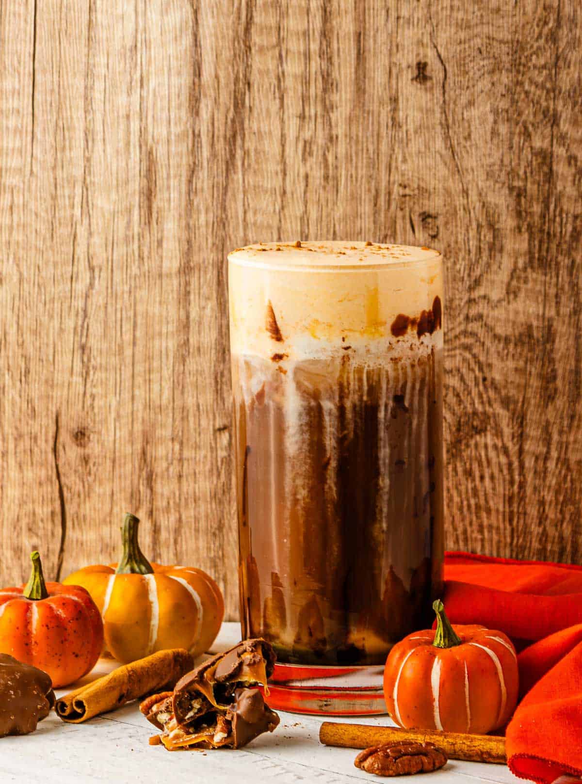 Iced pumpkin spice cold brew with cream, surrounded by small pumpkins, pecans, cinnamon sticks, and autumn decor.