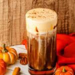 Tall glass of Pumpkin Cream Cold Brew with foam, surrounded by small pumpkins, cinnamon sticks, pecans, and a red cloth—a cozy Starbucks Copycat ready for autumn sipping.