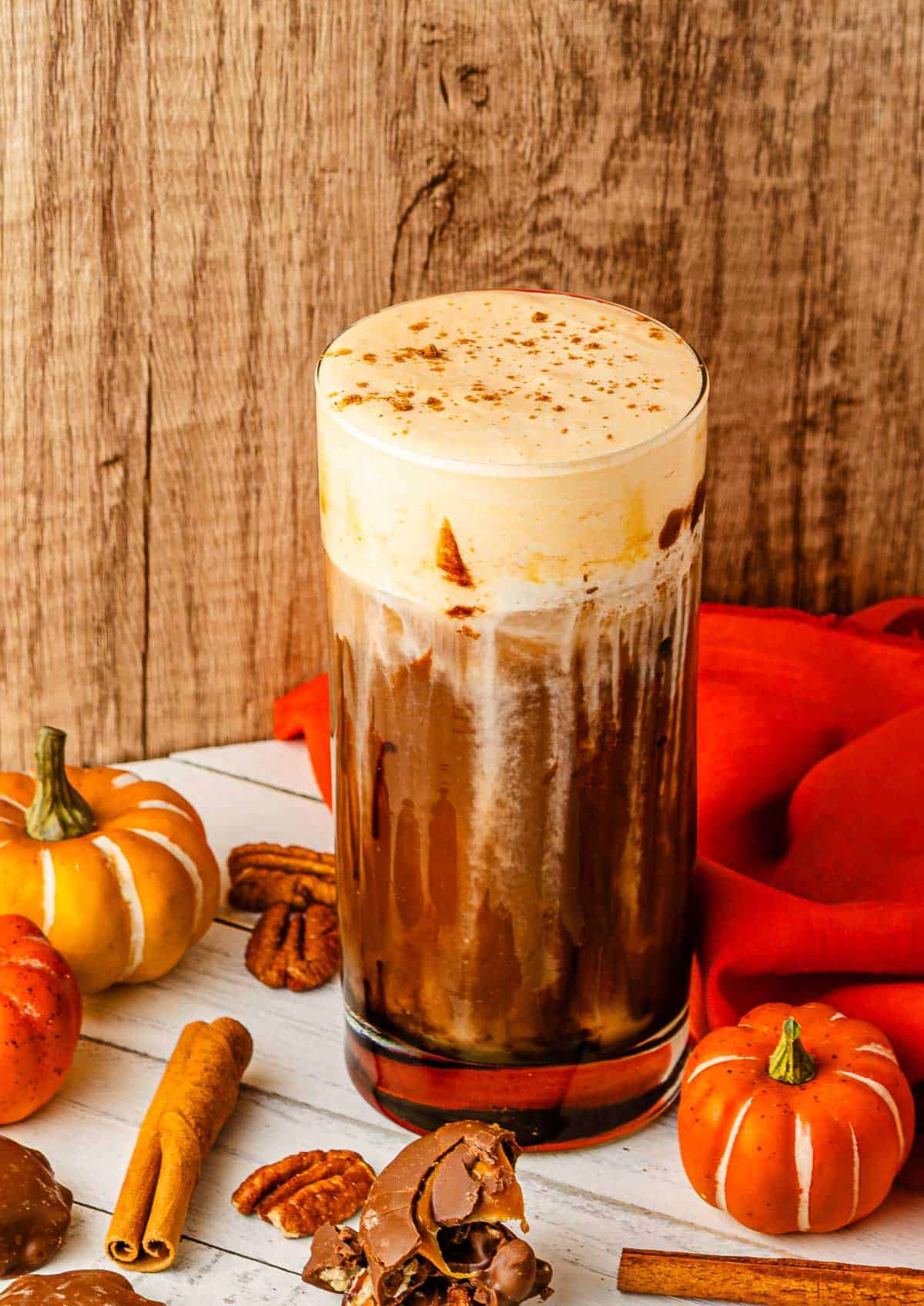Tall glass of Pumpkin Cream Cold Brew with foam, surrounded by small pumpkins, cinnamon sticks, pecans, and a red cloth&mdash;a cozy Starbucks Copycat ready for autumn sipping.