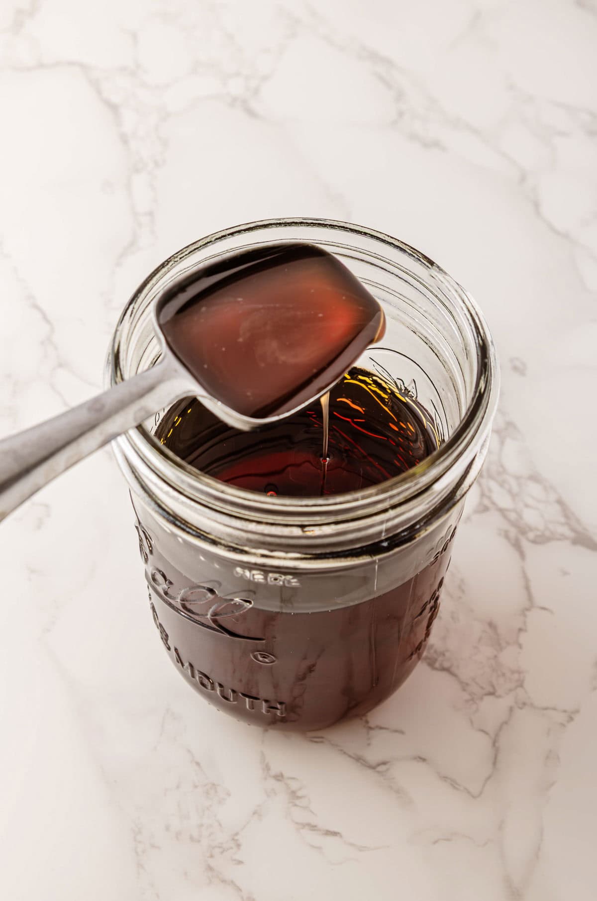 A spoon lifting dark Caramel Turtle syrup from a glass jar on a white marble surface.