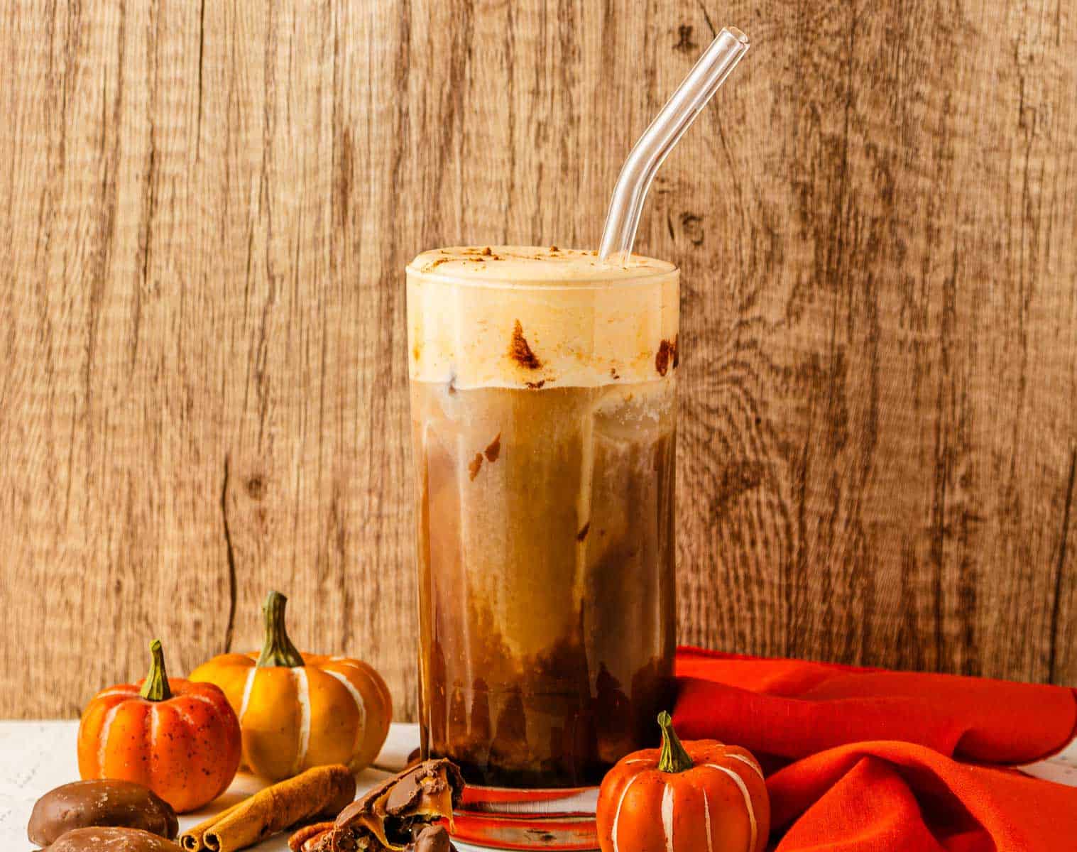 Iced cold brew with foam in a glass, surrounded by small decorative pumpkins and an orange cloth&mdash;perfect for pairing with pumpkin cream flavors.