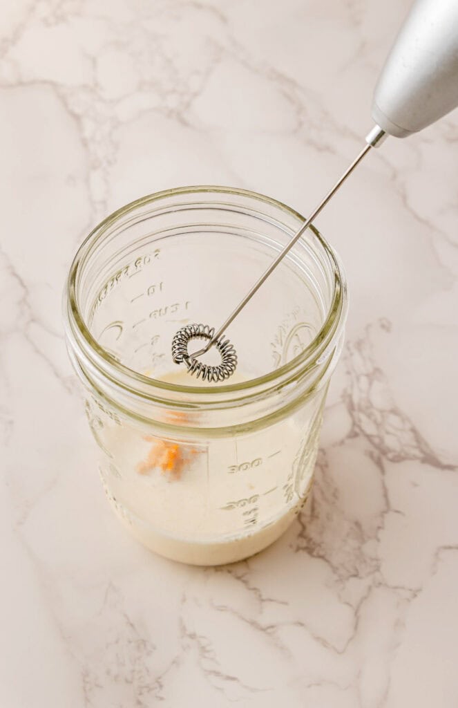 A handheld frother mixing Pumpkin Cream Cold Brew in a glass mason jar on a marble countertop, creating a delicious Starbucks Copycat treat.
