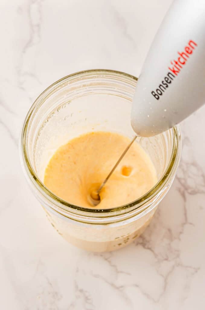 An immersion blender mixes a creamy yellow liquid in a glass jar on a white marble surface, creating a luscious Starbucks Copycat treat.