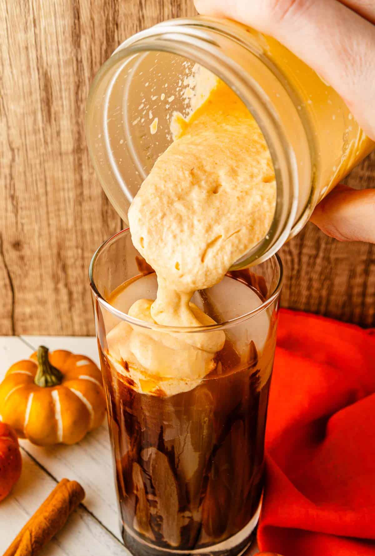 Pumpkin cream is being poured from a jar into a glass of iced coffee, creating a delicious Pumpkin Cream Cold Brew. A pumpkin and cinnamon stick sit nearby, making this drink the perfect Starbucks Copycat for fall.