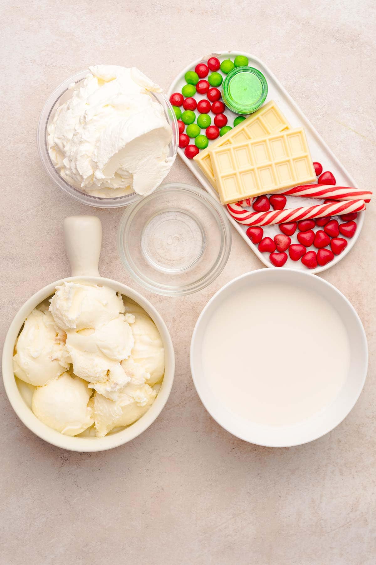 Bowls of vanilla ice cream, whipped cream, white chocolate, candies, candy canes, and a small bottle of extract&mdash;perfect ingredients for a festive Grinch Freakshake recipe.