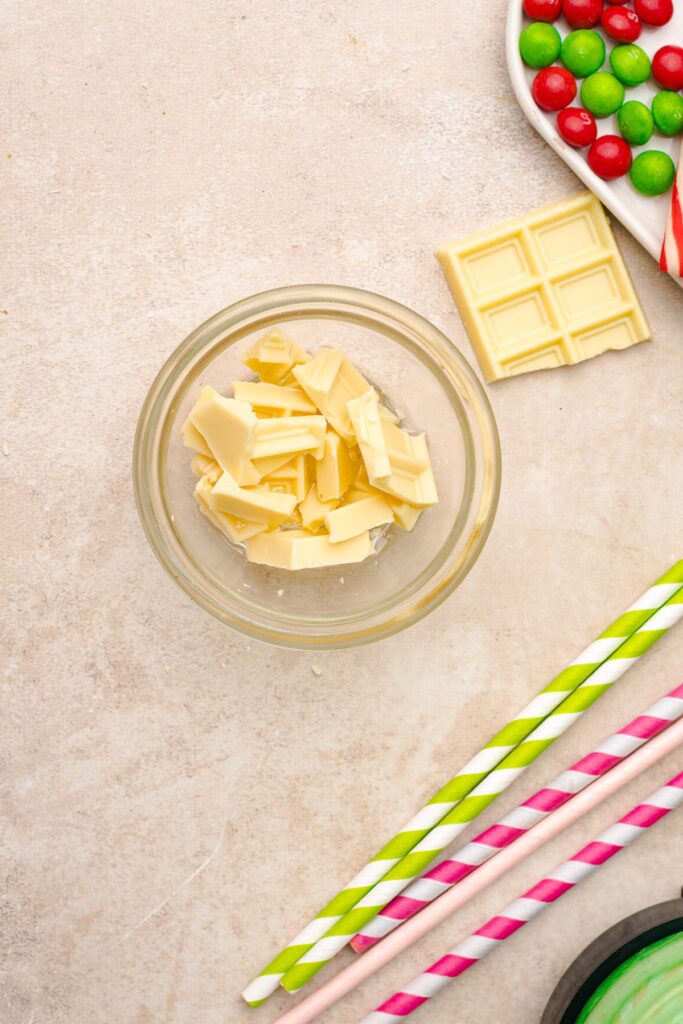 Bowl of white chocolate pieces, white chocolate bar, candy pieces, and colorful striped straws on a countertop&mdash;perfect for creating a Grinch Freakshake or trying out a new Freakshake Recipe.