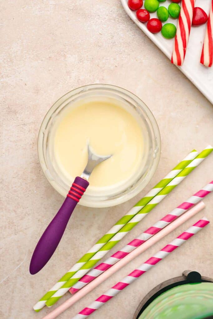Bowl of melted white chocolate with a spatula, colorful striped straws, candy canes, and candies nearby&mdash;perfect ingredients for a festive Grinch Freakshake recipe.