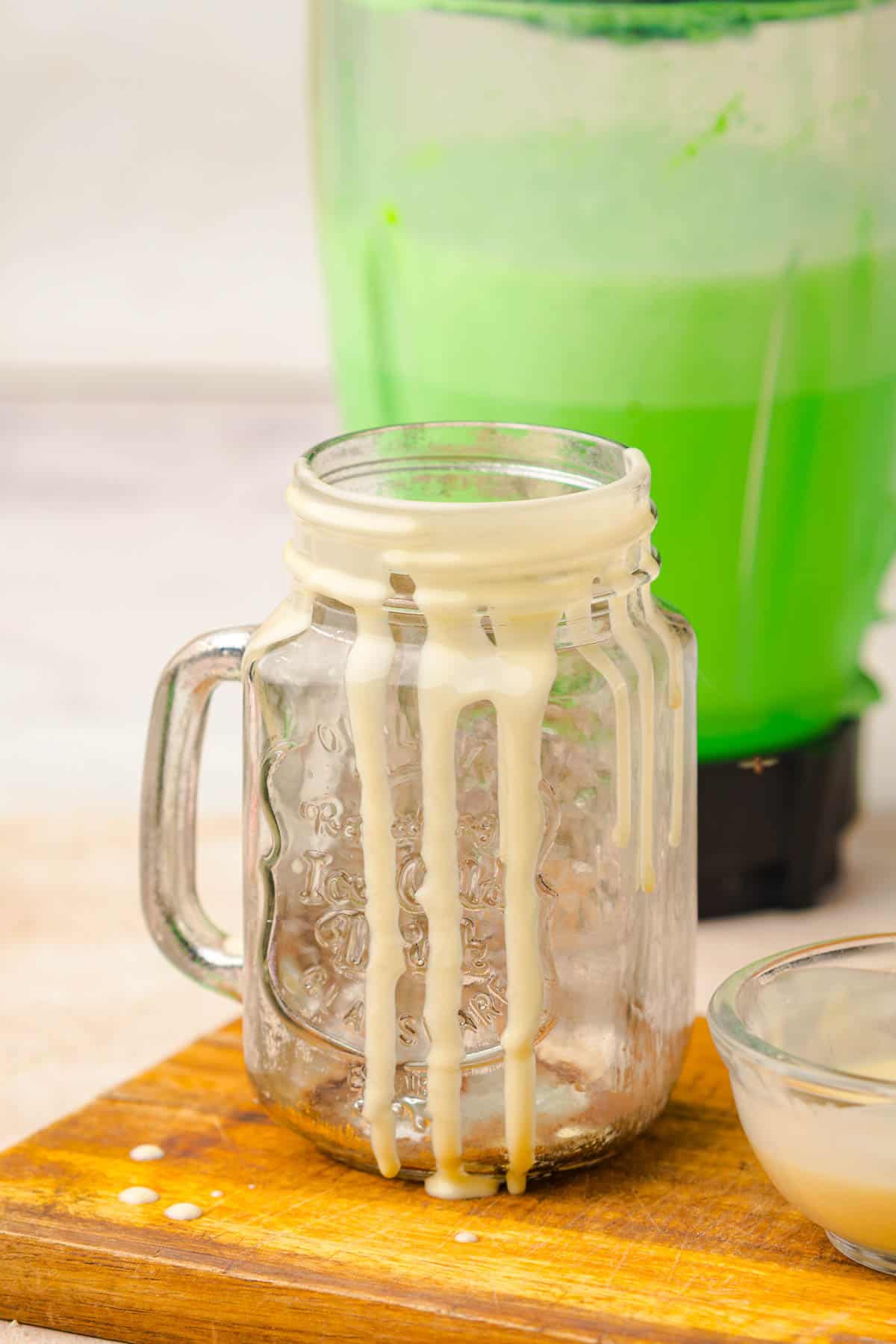 An empty mason jar with white sauce drips, a green smoothie in a blender, and a small bowl on a wooden board&mdash;perfect inspiration for your next Grinch Freakshake recipe.