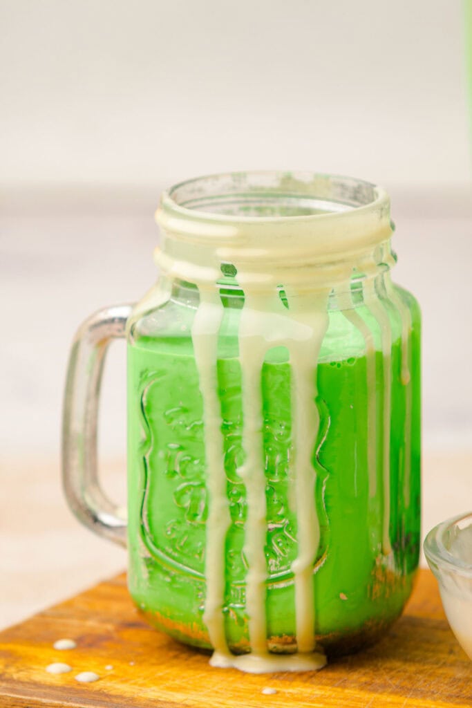 Mason jar with a green drink and creamy white drips on the sides, placed on a wooden surface&mdash;a perfect Grinch Freakshake to brighten your holiday festivities.