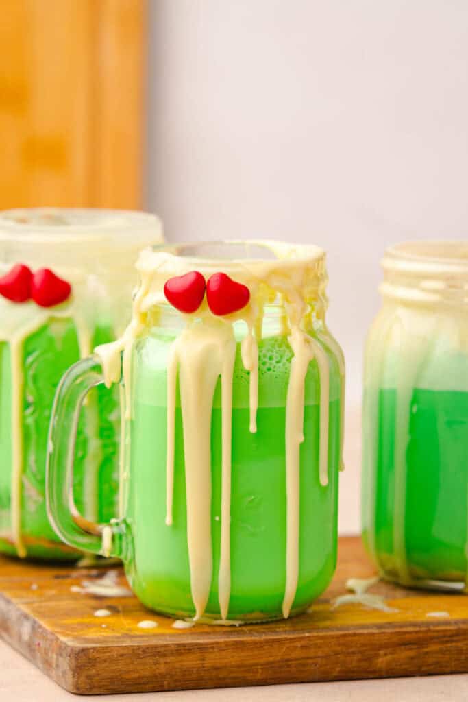 Three mason jars of bright green Grinch Freakshake drinks with dripping white chocolate and red candy hearts on the rim.