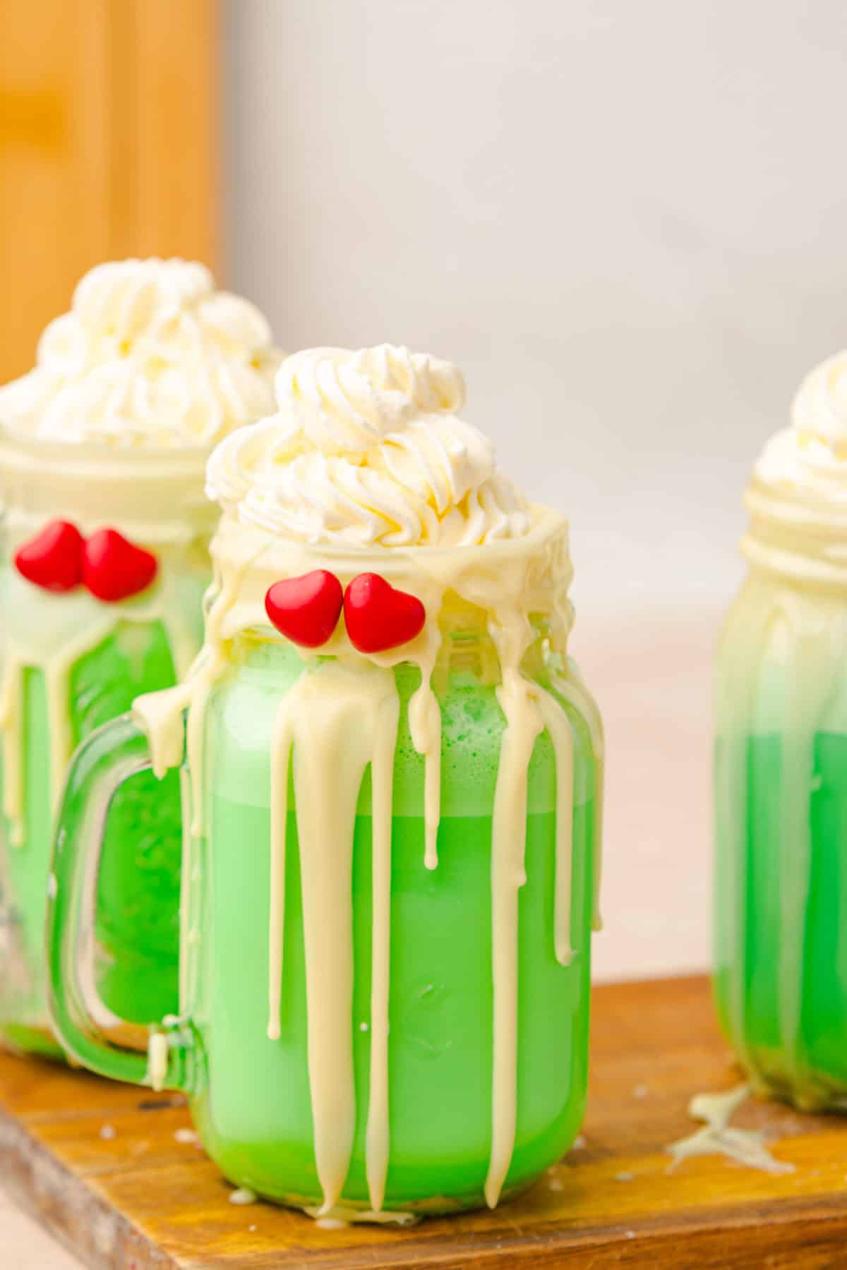Three green Grinch Freakshakes in jars with whipped cream, white chocolate drips, and red heart decorations are displayed on a wooden board.