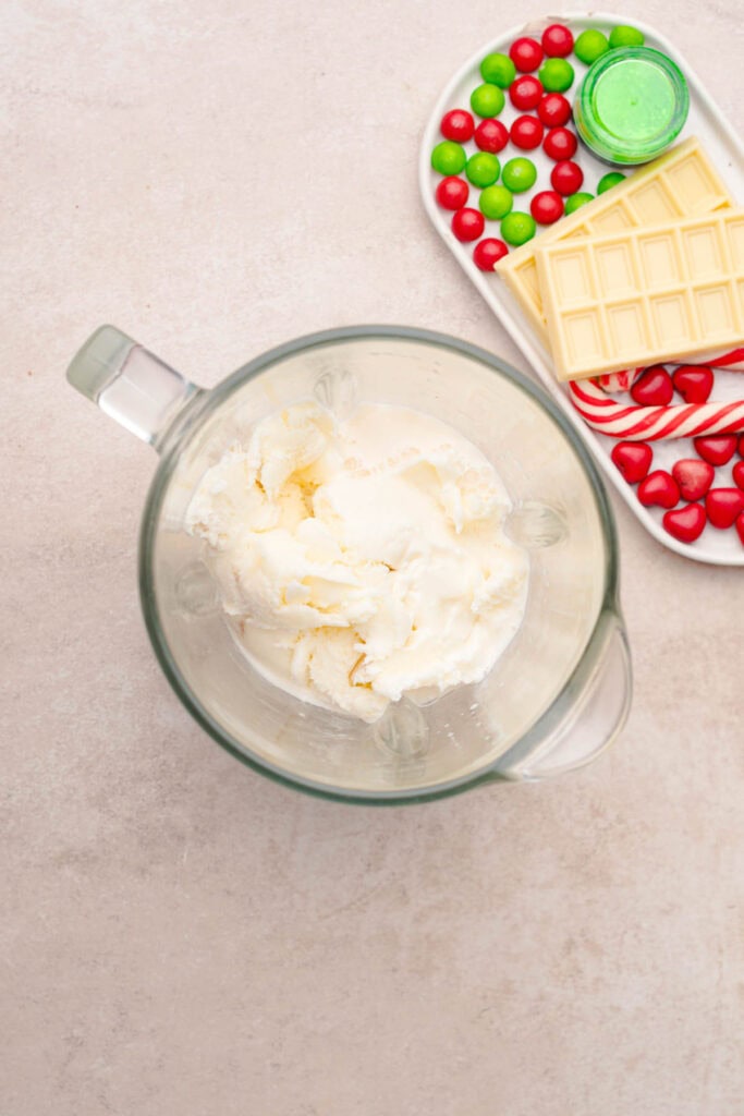 Glass blender filled with ice cream beside a tray of white chocolate, red and green candies, and green liquid&mdash;perfect for crafting a festive Grinch Freakshake recipe.