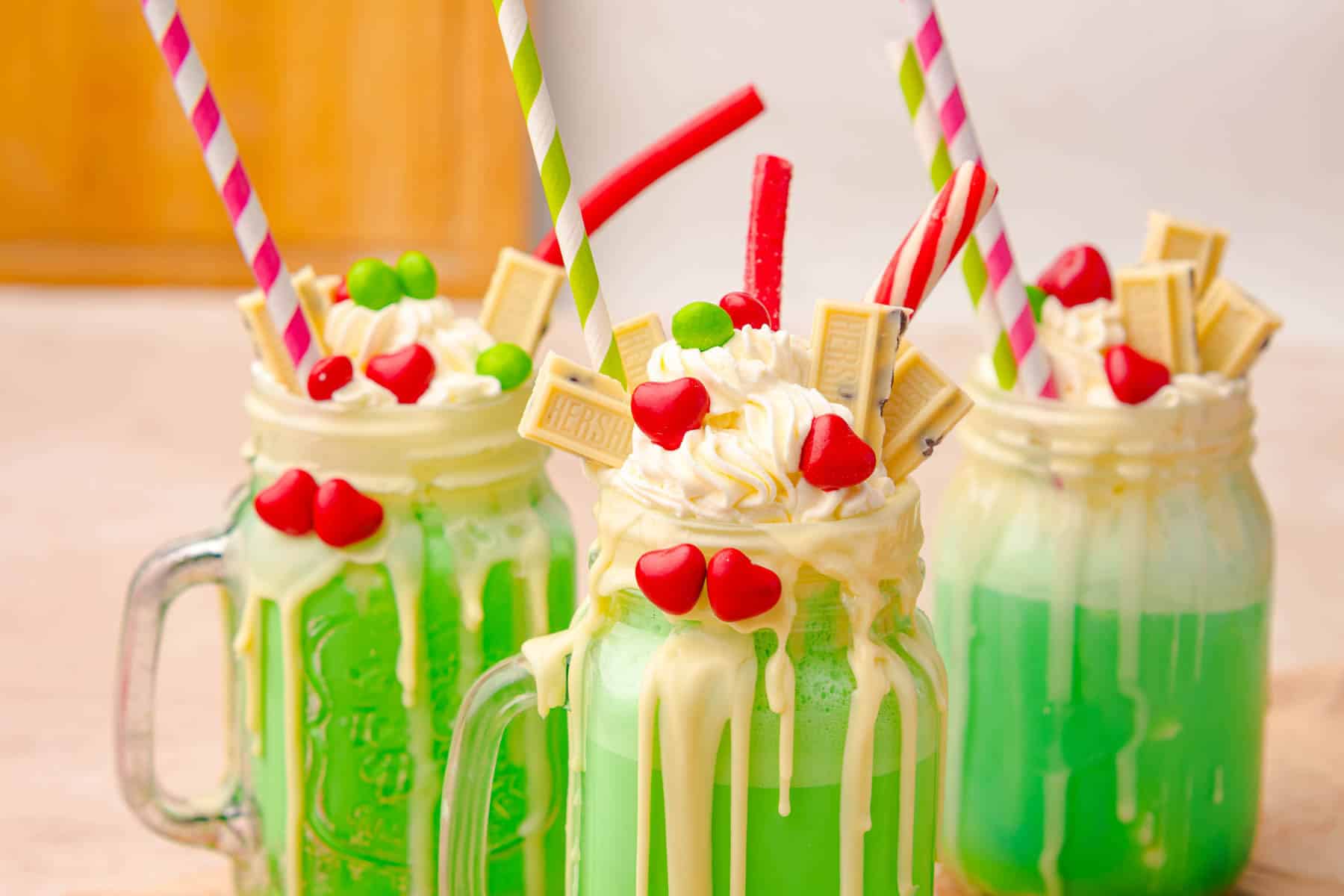 Three mason jar mugs filled with festive Grinch Freakshakes&mdash;green milkshakes, whipped cream, candy, and colorful straws. Try this playful Grinch Freakshake Recipe for a fun holiday treat!.