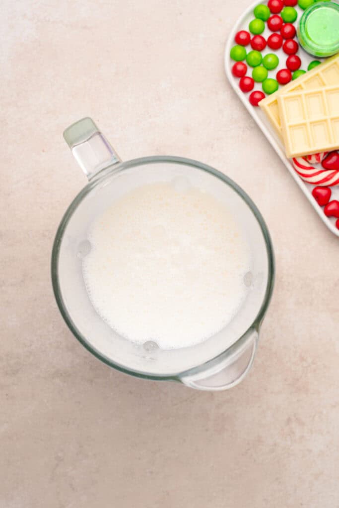 A glass measuring cup filled with frothy milk sits near a tray of colorful candies and white chocolate&mdash;perfect ingredients for a festive Grinch Freakshake.