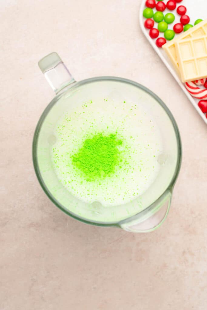 A glass measuring cup with milk and vibrant green matcha powder&mdash;perfect for crafting a festive Grinch Freakshake&mdash;sits beside a tray of candies and white chocolate.