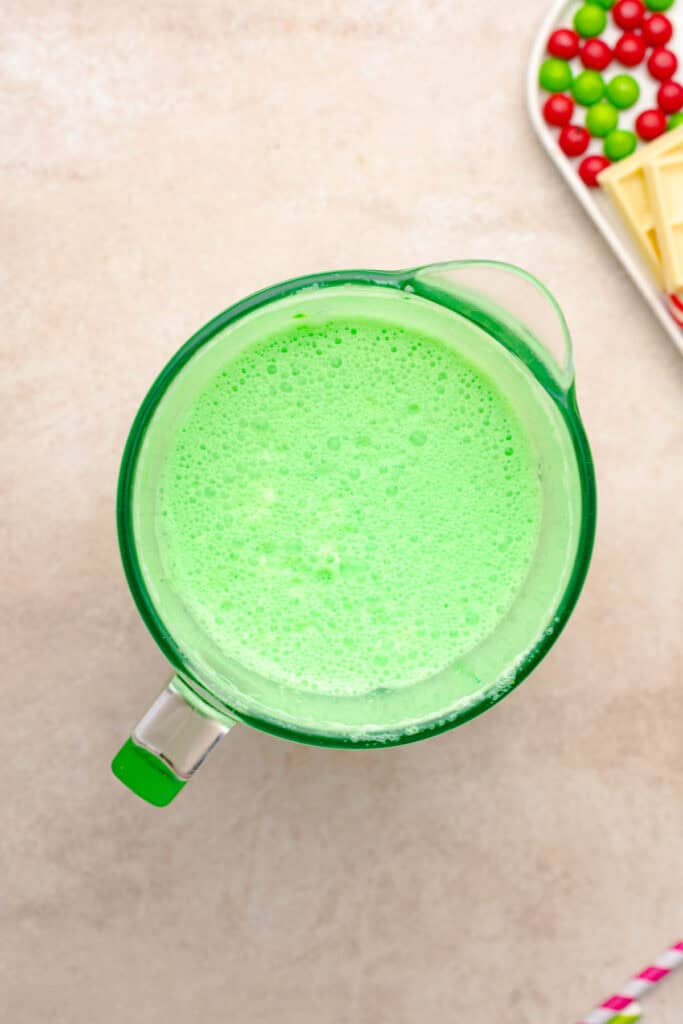 A glass pitcher filled with bright green frothy liquid, reminiscent of a Grinch Freakshake, sits beside a plate of red, green candies, and white chocolate.