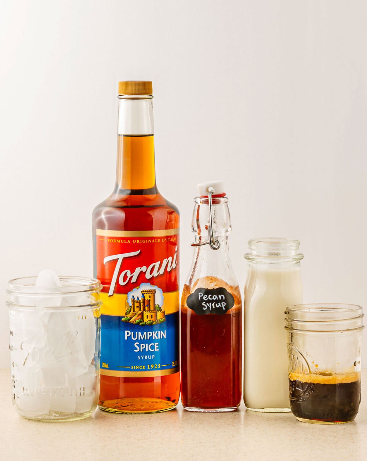 Pumpkin spice syrup, pecan syrup, milk, espresso, and a glass with ice are lined up on a counter for this Pumpkin Pecan Oatmilk Shaken Espresso&mdash;a perfect Starbucks Copycat treat at home.