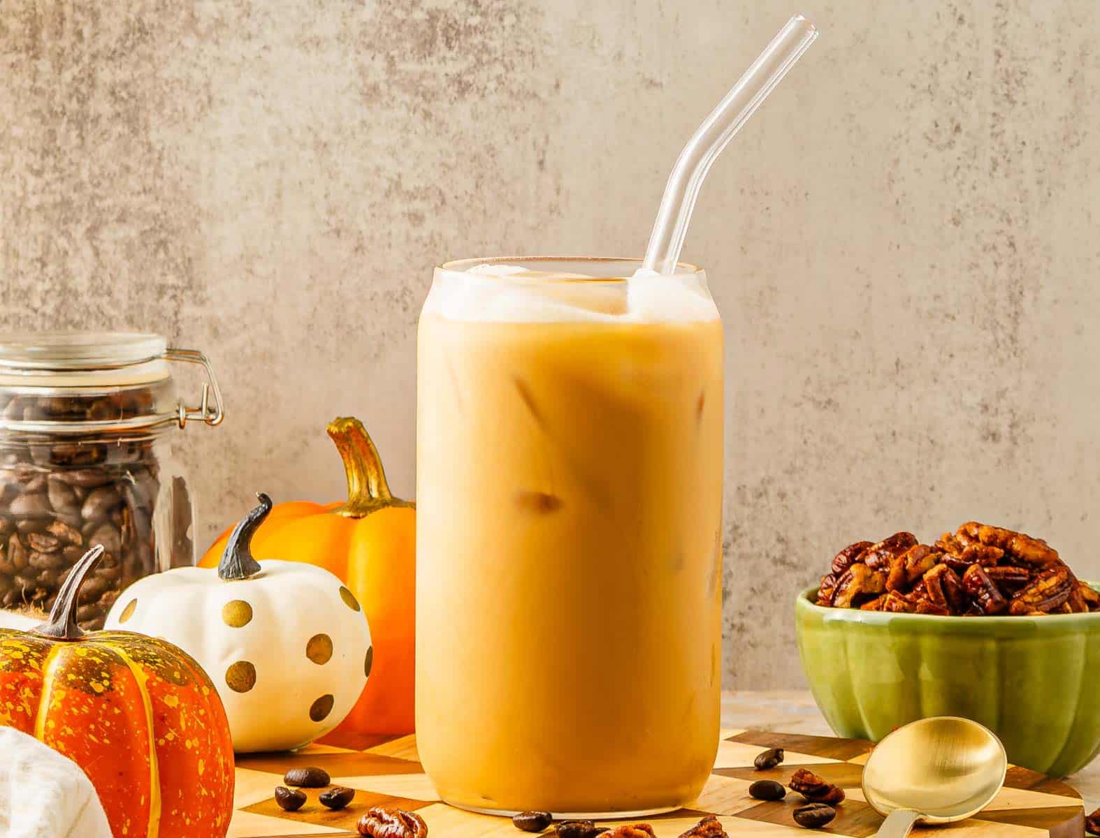 Iced pumpkin spice latte with a glass straw, surrounded by pumpkins, pecans, coffee beans, and a gold spoon&mdash;a true Pumpkin Pecan delight inspired by a Starbucks Copycat recipe.