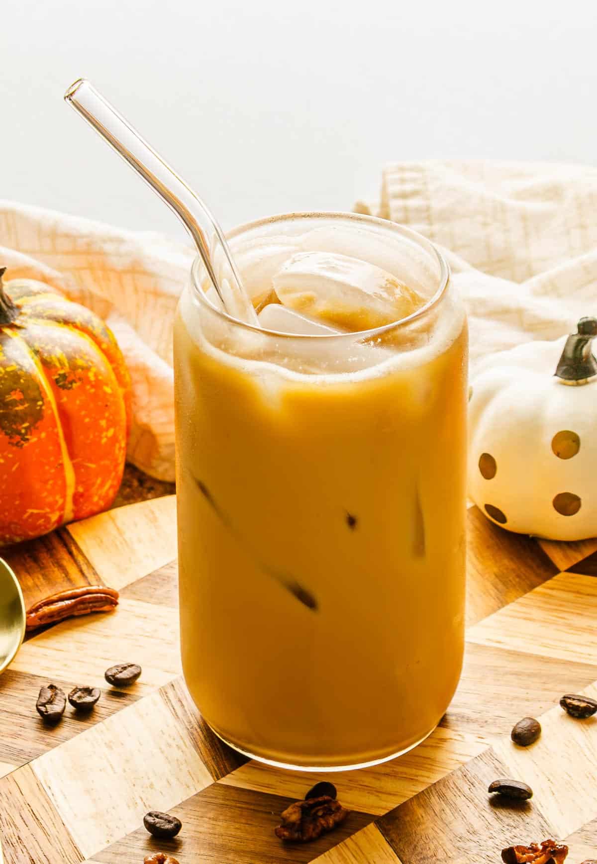 A glass of iced coffee with a straw, surrounded by coffee beans, pecans, and two small decorative pumpkins&mdash;perfect for recreating a Starbucks Copycat Pumpkin Pecan Oatmilk Shaken Espresso at home.