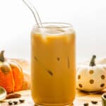 A glass of iced Pumpkin Pecan Oatmilk Shaken Espresso with a glass straw, surrounded by pumpkins and pecans on a wooden surface, captures the cozy vibes of fall in this Starbucks Copycat recipe.
