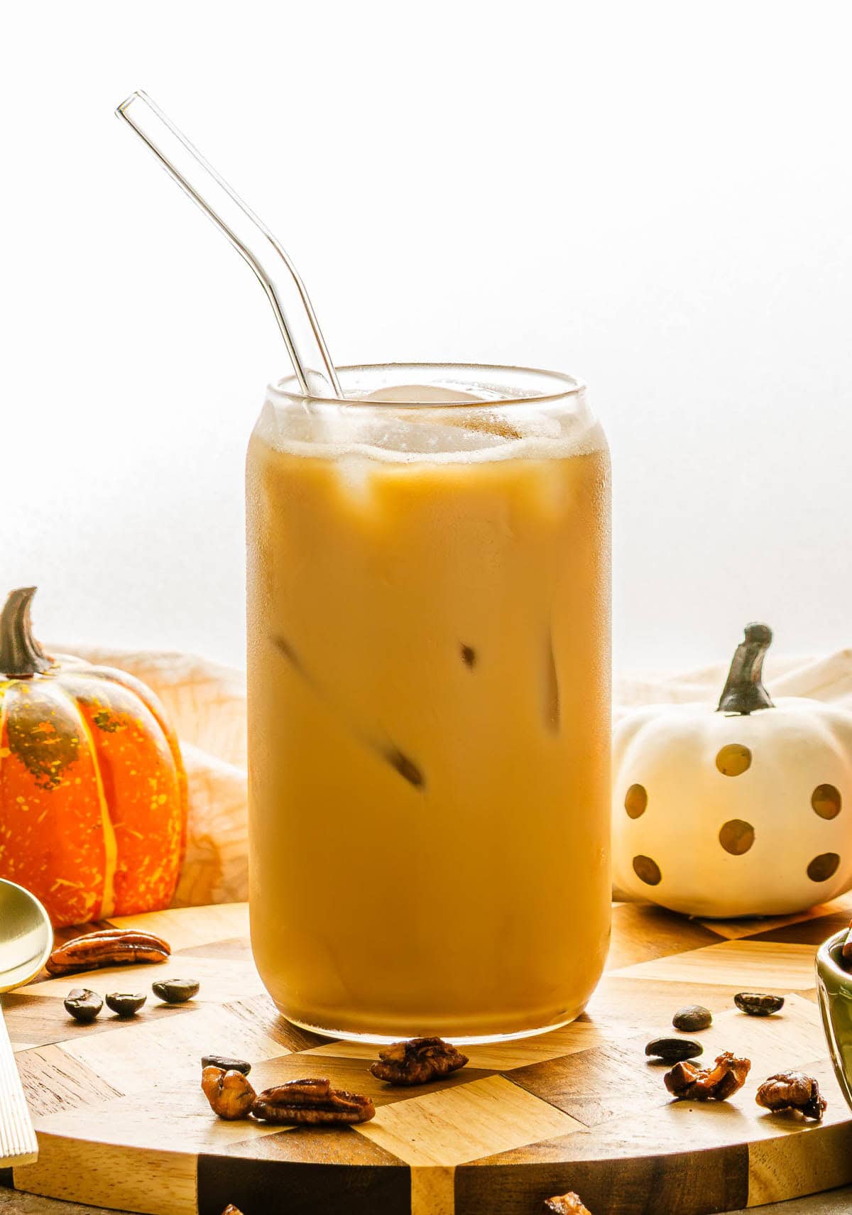 A glass of iced Pumpkin Pecan Oatmilk Shaken Espresso with a glass straw, surrounded by pumpkins and pecans on a wooden surface, captures the cozy vibes of fall in this Starbucks Copycat recipe.