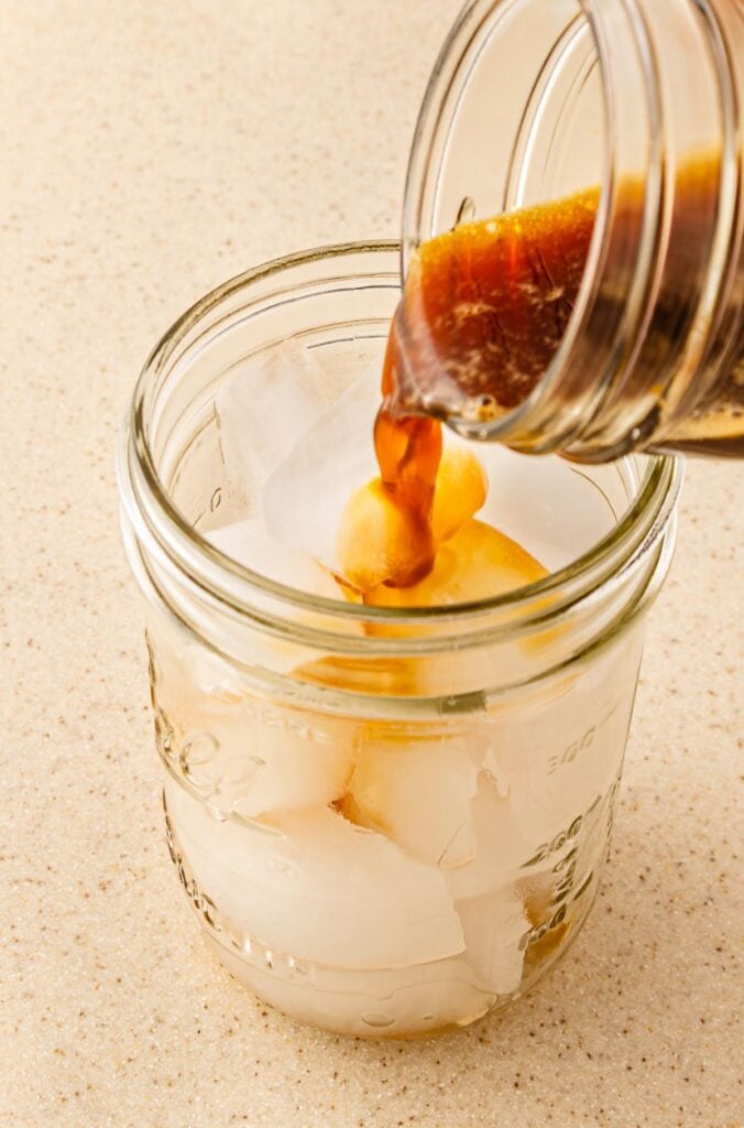 Iced coffee being poured into a mason jar filled with ice cubes on a light countertop, inspired by the popular Starbucks Copycat Oatmilk Shaken Espresso.