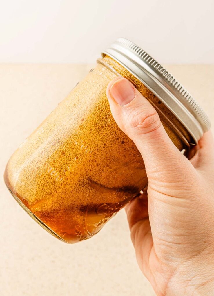 A hand holding a mason jar filled with foamy brown liquid against a light background, showcasing a Pumpkin Pecan Oatmilk Shaken Espresso&mdash;your favorite Starbucks copycat recipe at home.