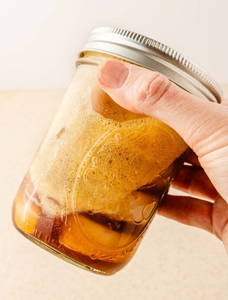 A hand holding a glass jar filled with foamy brown liquid, sealed with a metal lid&mdash;just like a homemade Starbucks Copycat Oatmilk Shaken Espresso.