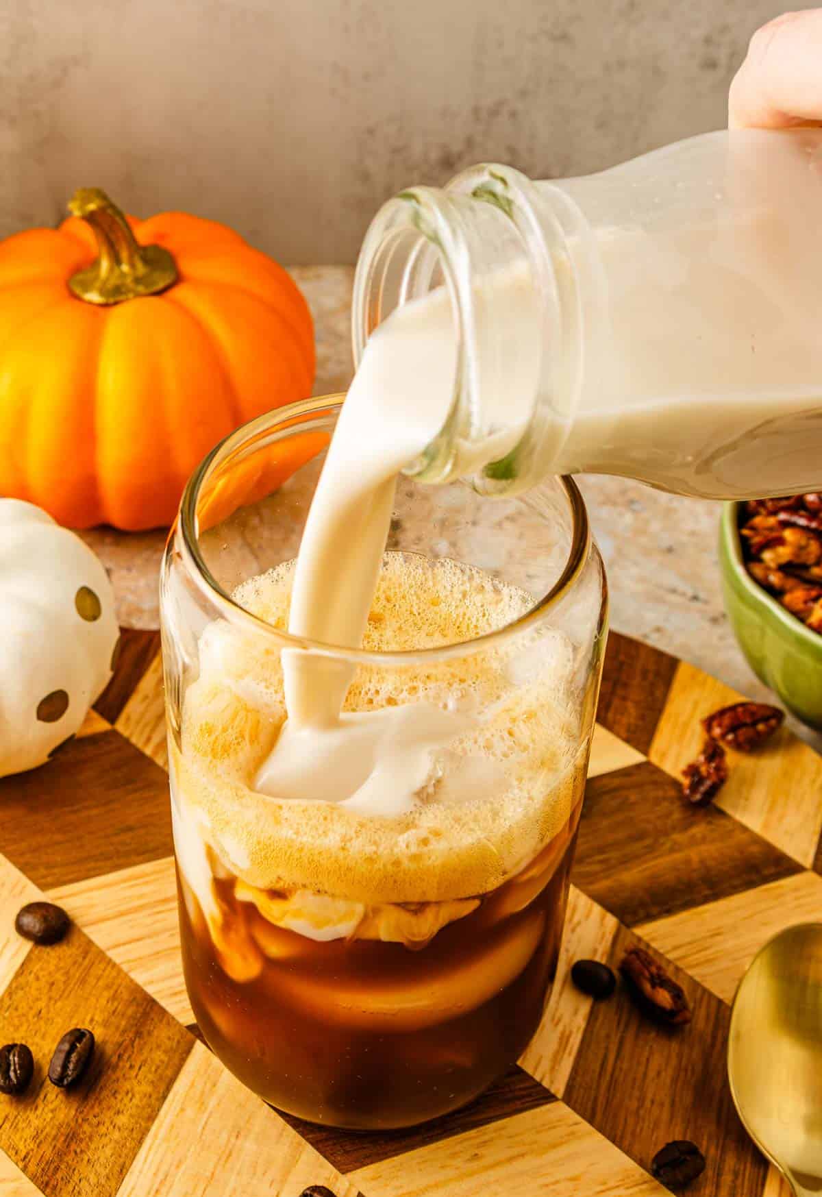 Milk being poured into iced coffee in a glass, inspired by a Starbucks Copycat Pumpkin Pecan Oatmilk Shaken Espresso, with a small pumpkin and coffee beans nearby on a wooden board.
