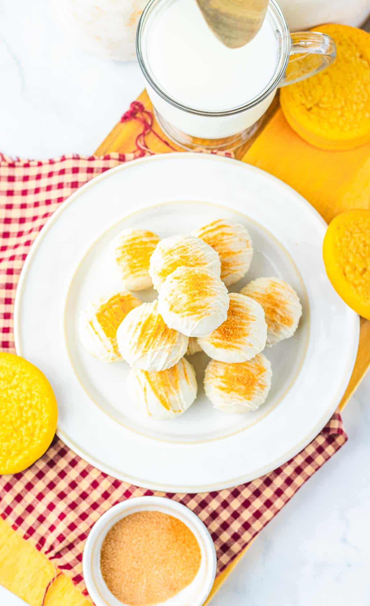 A plate of round, white snickerdoodle cookies with yellow sugar, next to milk and yellow cookies on a gingham cloth&mdash;a delightful recipe for any occasion.