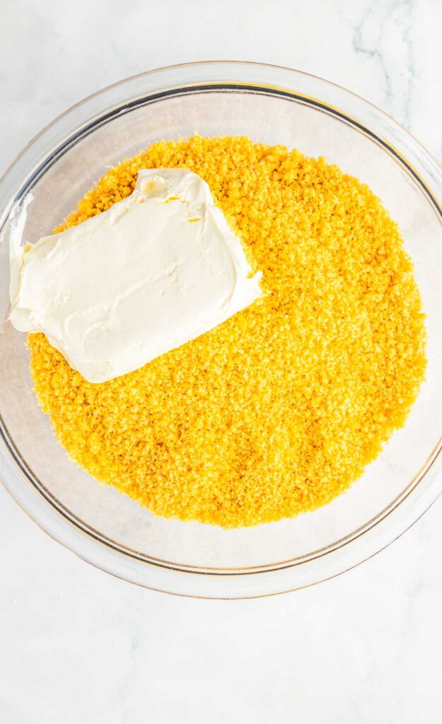 A glass bowl with crushed yellow graham crackers and a block of cream cheese on top&mdash;perfect for crafting a delicious Snickerdoodle truffles recipe.