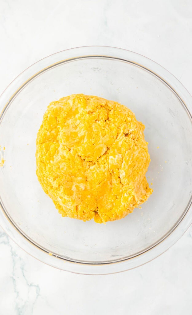 A ball of yellow dough for Snickerdoodle Truffles rests in a clear glass bowl on a white marble surface.