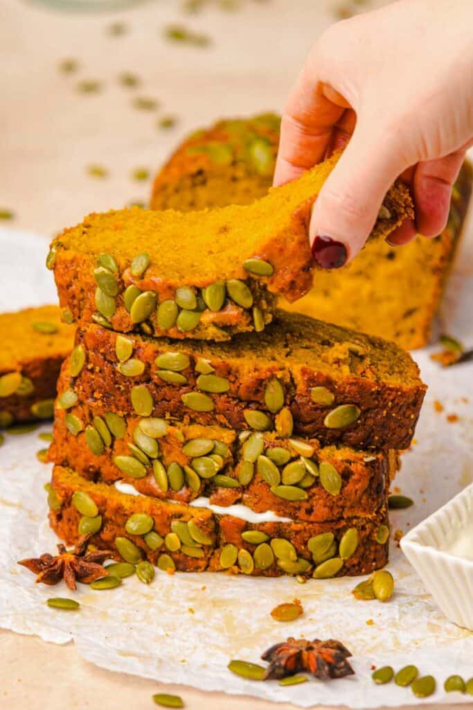 Hand lifting a slice of pumpkin bread topped with pumpkin seeds from a stack on parchment paper-a delicious Starbucks copycat treat.