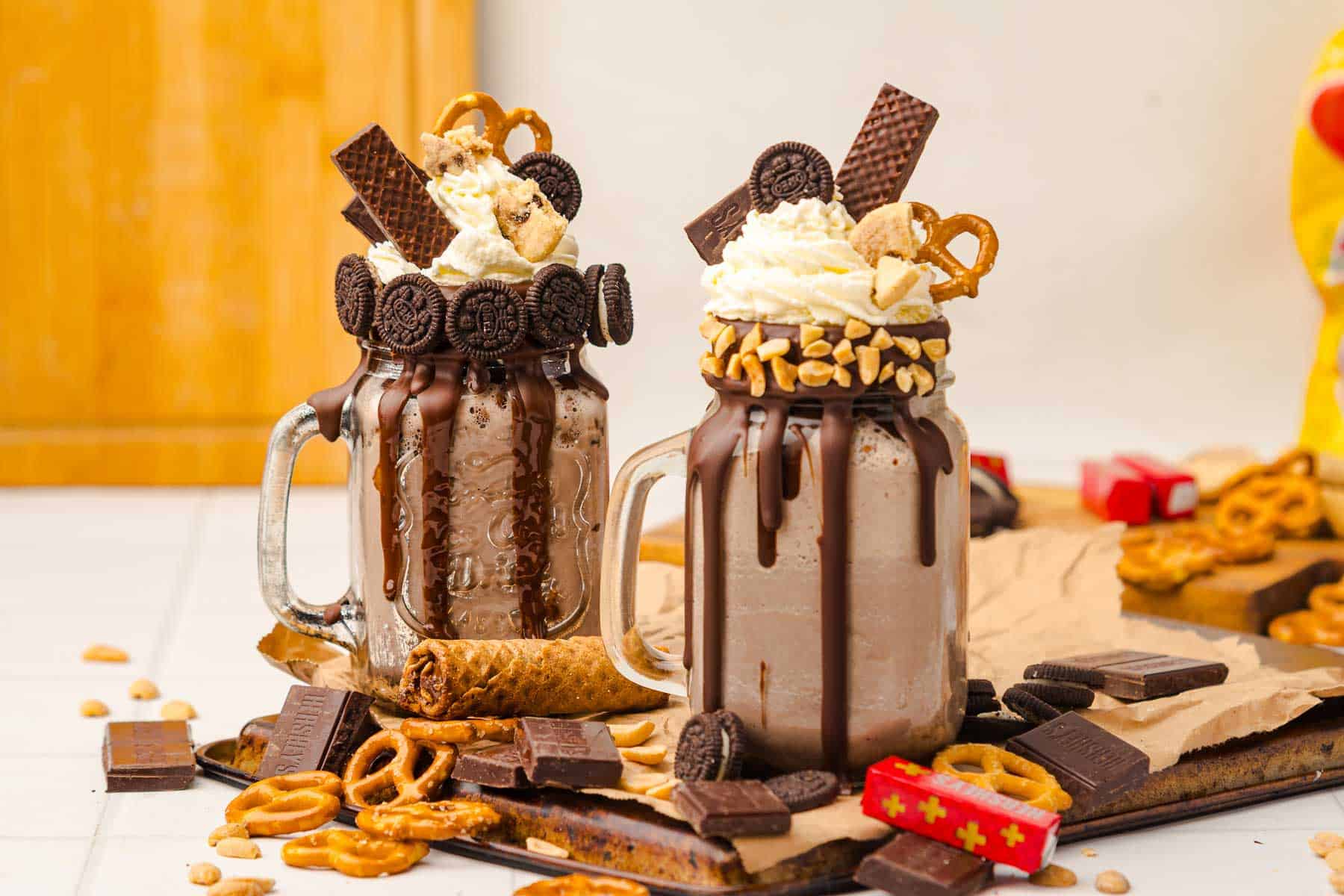 Two chocolate milkshakes in mason jars, topped with whipped cream, cookies, wafers, peanuts, and pretzels&mdash;a decadent Chocolate Freakshake Recipe perfect for satisfying your sweet cravings.