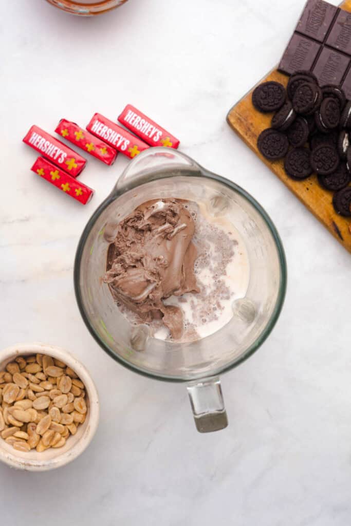 Chocolate ice cream and milk in a blender, with chocolate bars, cookies, and peanuts nearby on a white surface, perfect for creating a delicious Chocolate Freakshake Recipe.
