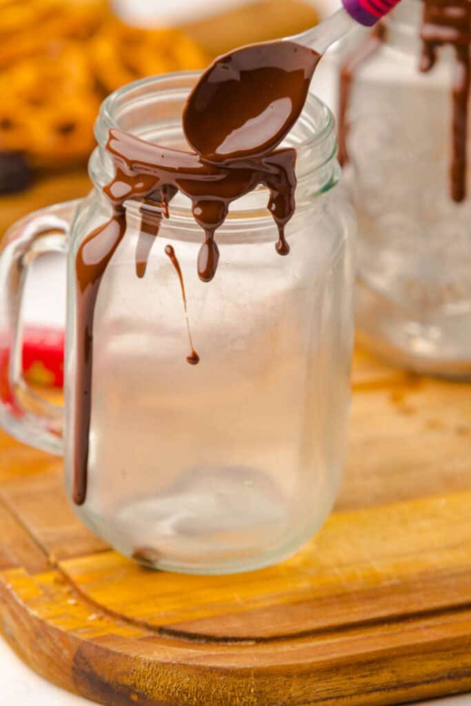 A spoon drizzles melted chocolate on the rim of an empty mason jar, setting the stage for a delicious Chocolate Freakshake Recipe on a wooden board.