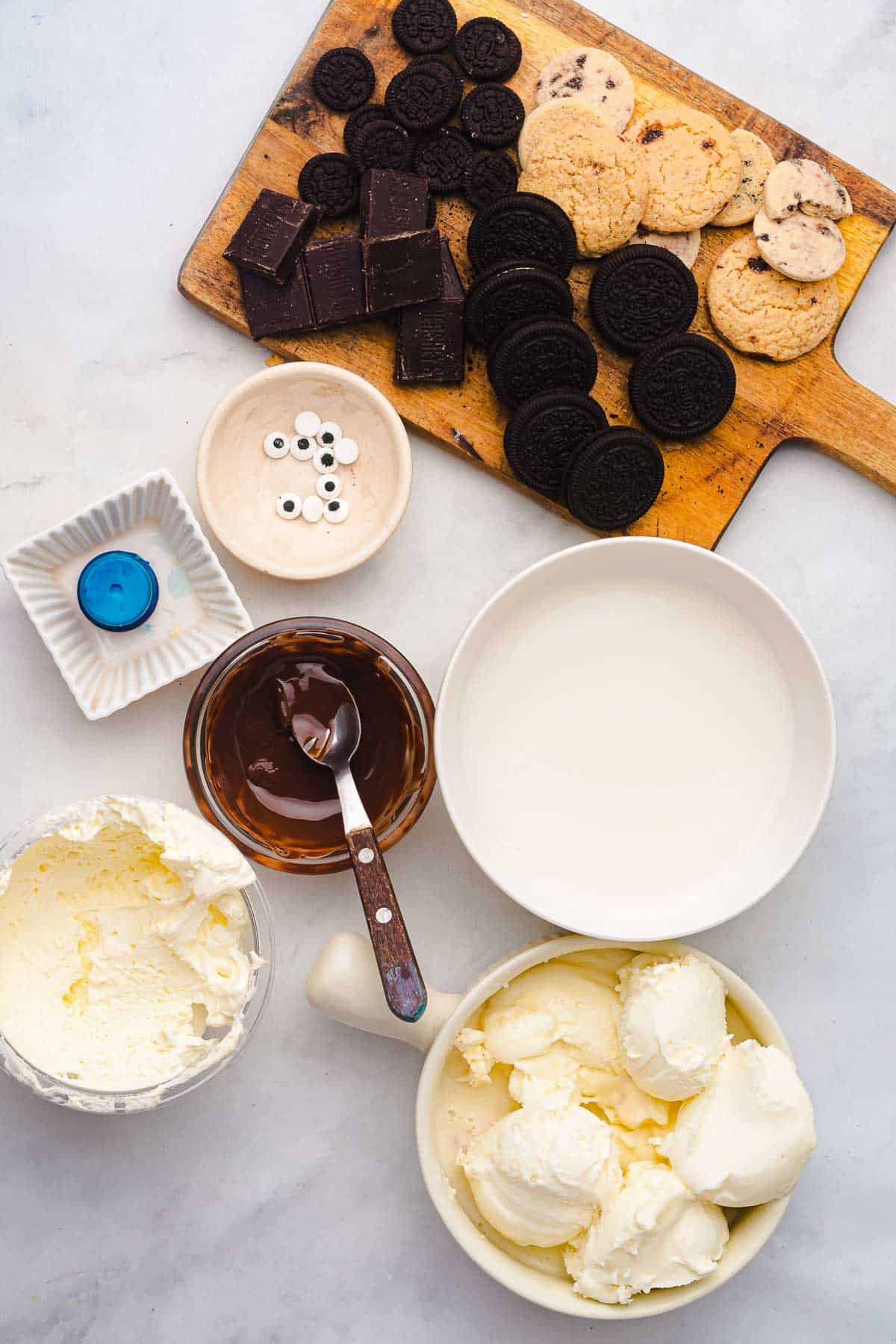 Assorted cookies, chocolate, cream, ice cream, candy eyes, and blue food coloring on a white surface&mdash;perfect for creating a fun Cookie Monster Milkshake.