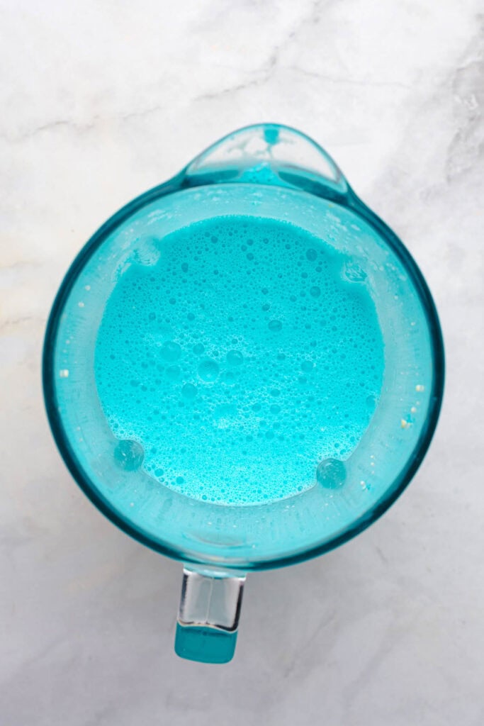 A blender filled with a bright blue frothy liquid, viewed from above on a light marble surface&mdash;perfect for whipping up a fun Cookie Monster milkshake recipe.
