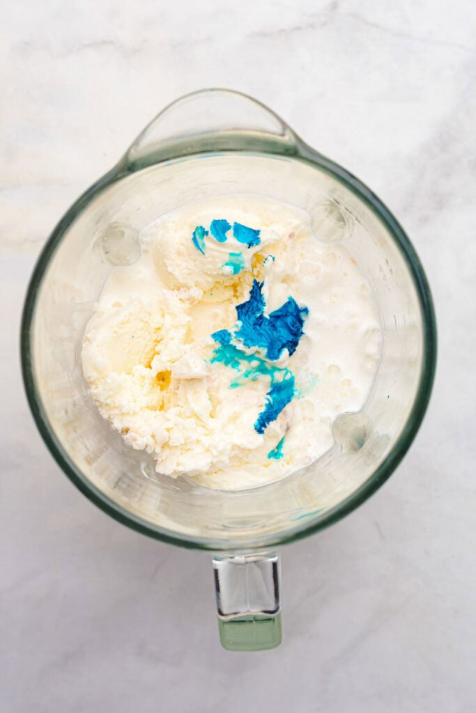 A blender with vanilla ice cream, milk, and blue food coloring on a marble surface, ready to whip up a fun Cookie Monster milkshake.