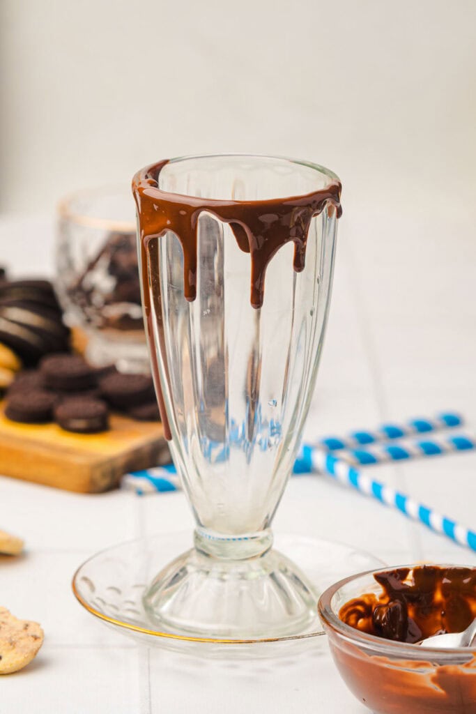 Empty glass sundae cup with chocolate dripping from the rim, surrounded by cookies and blue striped straws&mdash;a tempting setup for a perfect Cookie Monster Milkshake.