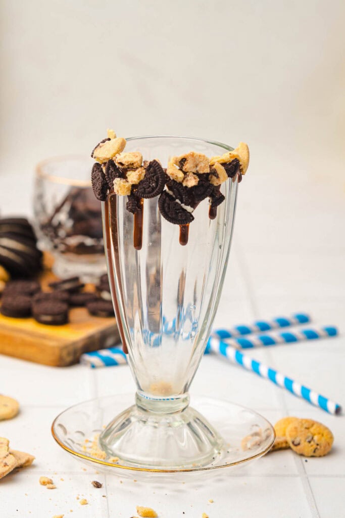 Empty milkshake glass with cookie pieces and chocolate on the rim, with cookies and straws in the background&mdash;perfect inspiration for your next Cookie Monster milkshake or creative milkshake recipe.