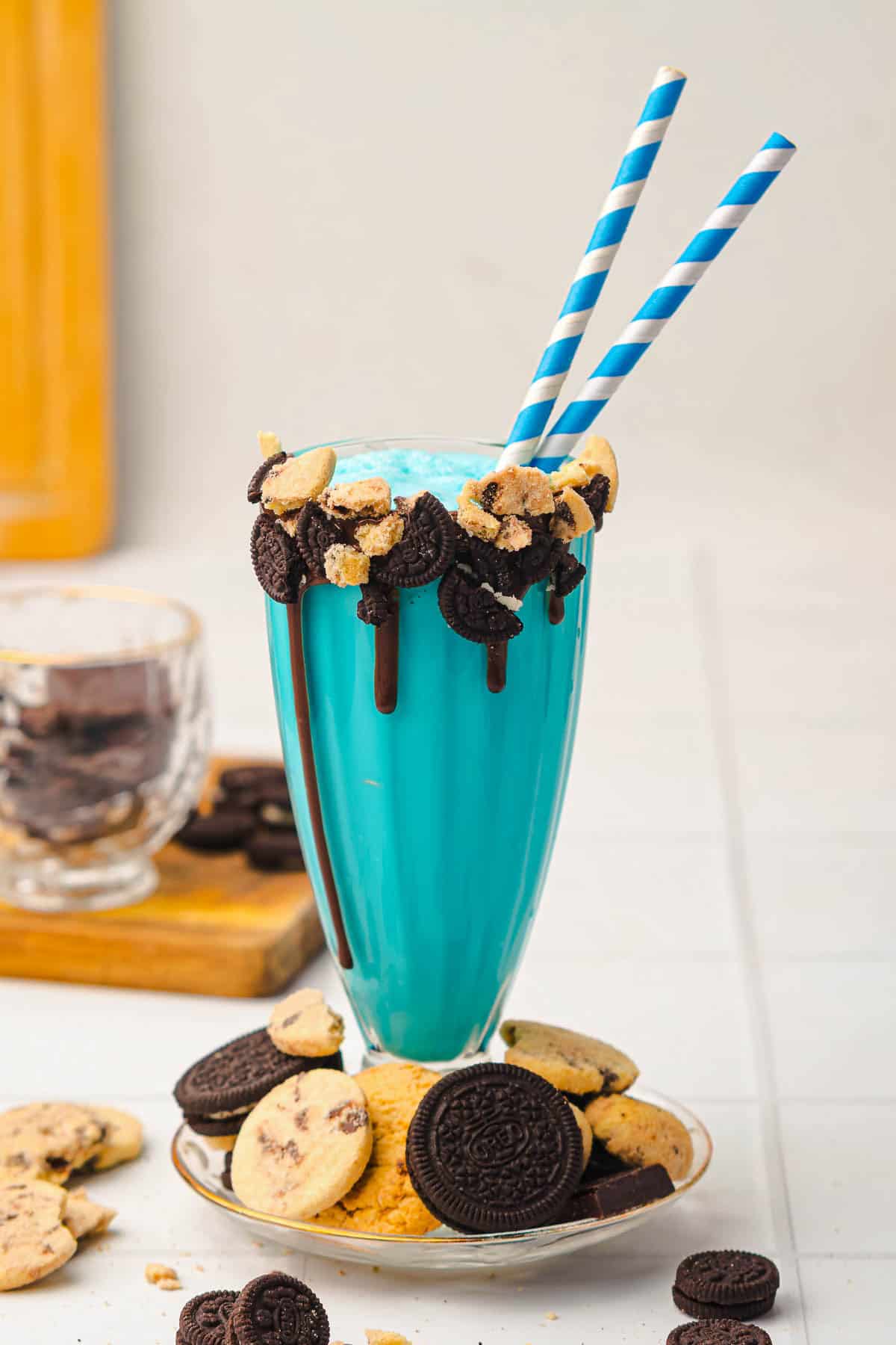 Indulge in this Cookie Monster Milkshake: a vibrant blue milkshake in a tall glass with blue-striped straws, topped and surrounded by cookies&mdash;a fun and delicious Cookie Monster Recipe perfect for any sweet craving.