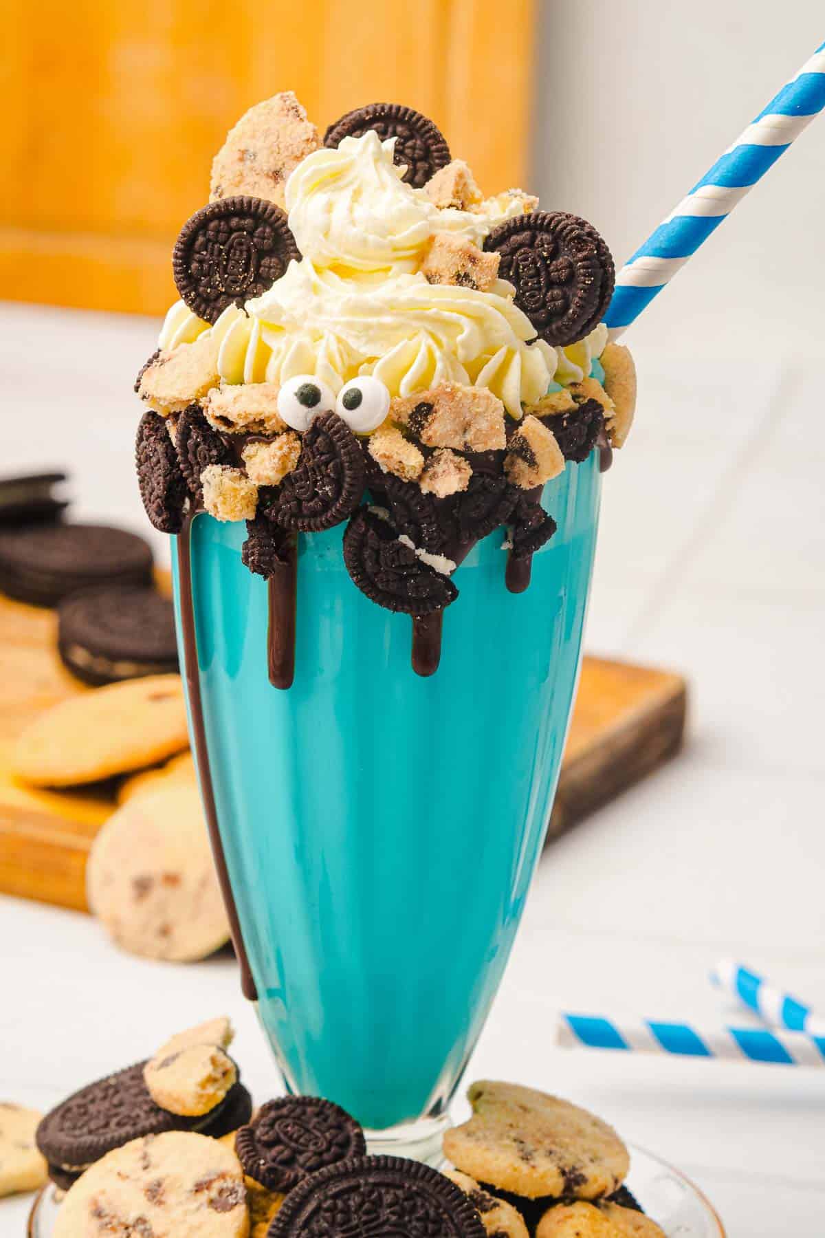 Blue Cookie Monster milkshake topped with whipped cream, mini cookies, chocolate, and candy eyes, served with a blue-striped straw&mdash;an irresistible Cookie Monster dessert!.