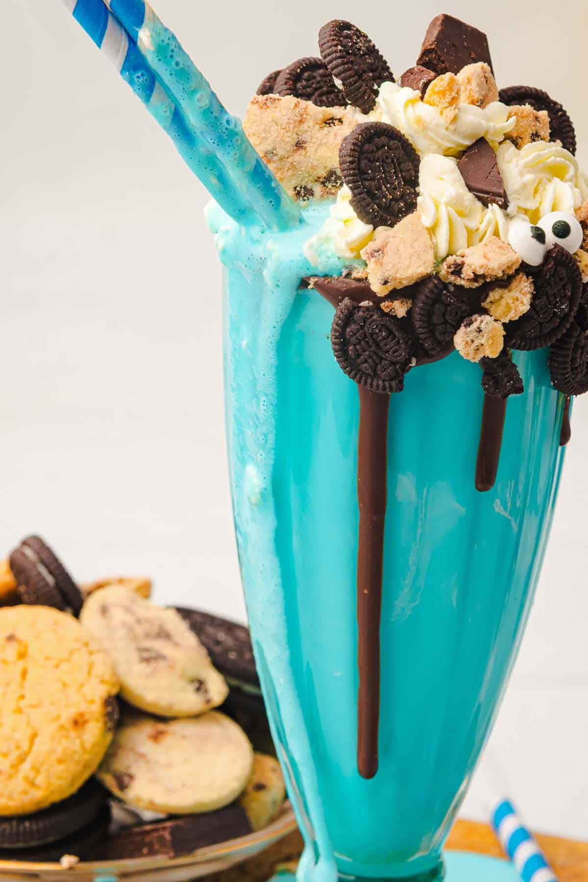 A Cookie Monster Milkshake topped with whipped cream, cookies, chocolate, and two striped straws, this blue milkshake is the perfect treat for any dessert lover.